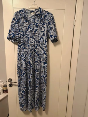 Blåvit långklänning från H&M - Säljer en blåvit långklänning från H&M i storlek M. Klänningen har ett tropiskt bladmönster, korta ärmar, krage och knappar framtill. Tillverkad i mjuk och sval viskos, perfekt för varma dagar. Snygg och bekväm passform som ger en avslappnad look.