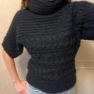 Svart stickad polotröja med kort ärm - Mysig svart stickad polotröja med grov kabelstickning och korta ärmar. Tröjan har en hög polokrage och ett chunky mönster som ger en cool och avslappnad vibe. Storlek S, normal i storleken. Tröjan är i bland annat 30% mohair och 15% ull. 