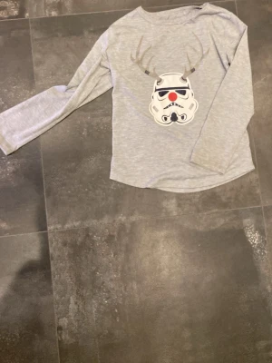 Grå Star Wars långärmad tröja GAP - Grå långärmad tröja från GAP x Star Wars med ett tryck av en stormtrooper med renhorn och röd nos på bröstet. Perfekt för dig som älskar nördiga jultröjor och vill sticka ut lite extra under vintern. Mjuk och skön bomullsblandning.