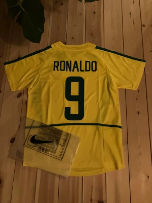 Nike Brasil Ronaldo #9 fotbollströja - Säljer en ikonisk Brasilien fotbollströja från Nike med Ronaldo och nummer 9 på ryggen. Tröjan är gul med gröna detaljer, officiell Brasil-logga och Nike Swoosh på bröstet. Tillverkad i polyester, perfekt för dig som älskar fotboll och klassiska landslagströjor. Inte riktig men jättebra kvalite