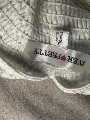 Vit långklänning från Ever Pretty - Säljer en vit långklänning från Ever Pretty med spets upptill och korta ärmar. Klänningen har v-ringad rygg, smock i midjan och ett medföljande bälte. Materialet är mjukt och lätt, med spetsdetaljer som ger en romantisk känsla.