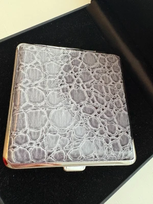 Grå plånbok med reptilmönster - Snygg grå plånbok med reptilmönstrad yta och silverfärgad metallram. Plånboken har ett stilrent och kompakt format med elastiska band på insidan för att hålla kort eller sedlar på plats. Perfekt för dig som gillar en unik och modern look.