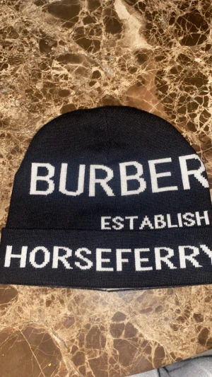 Burberry mössa med Horseferry-tryck - Snygg svart mössa från Burberry med vit text 'BURBERRY ESTABLISH HORSEFERRY' framtill. Mössan är stickad i mjuk bomull och har en klassisk form som passar perfekt för kalla dagar. Ikonisk design med tydlig logga och cool streetkänsla.