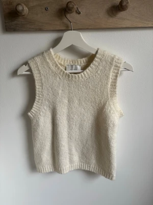 Stickad ärmlös tröja från Neo Noir - Stickad, ärmlös tröja från Neo Noir i mjuk och fluffig ullblandning. Färgen är ljus beige och modellen har rund hals med ribbade kanter. Materialet består av ull, polyester, akryl, alpaka och elastan. Perfekt att styla över en skjorta eller t-shirt.