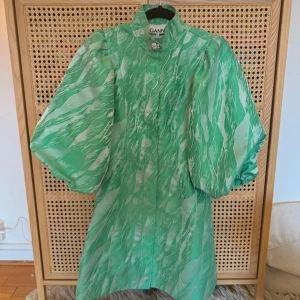 Grön klänning från Ganni - Säljer en superfin grön klänning från Ganni med unikt mönster i ljusa toner. Klänningen har puffiga långärmar, lite högre krage med dekorativ brosch och är tillverkad i glansigt polyester-material. Perfekt för dig som gillar statement-plagg och coola detaljer. Aldrig använd då den är för liten för mig. Nypris:2500kr