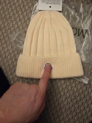 99 - Bilden visar en ribbstickad mössa i ljus beige med ett märke framtill