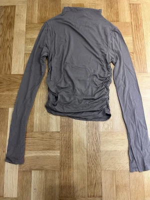 Långärmad polotopp - Säljer en brun/grå långärmad polotopp med scrunch på sidorna. Toppen har en hög krage och är tillverkad i mjukt och stretchigt material som sitter skönt mot kroppen. Perfekt att styla med jeans eller kjol för en cool look. Endast använd en gång 