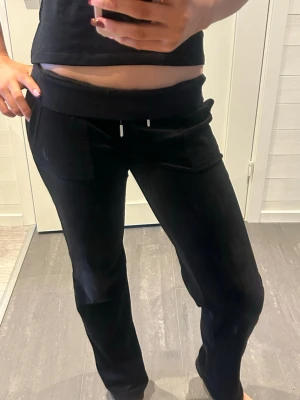 Svarta Juicy Couture mjukisbyxor - Svarta mjukisbyxor från Juicy Couture i mjuk velour med låg midja och raka ben. Byxorna har snörning i midjan, framfickor och bakfickor med knappdetalj i metall med Juicy-logga. Perfekta för en chill och avslappnad stil.