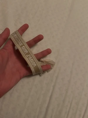 Beige Christian Dior J'Adior armband - Säljer ett beige Christian Dior J'Adior armband med broderad text och dekorativa tofsar i ändarna. Armbandet är justerbart och har en stilren, elegant look som passar perfekt till din outfit.