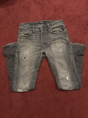 Grå slitna jeans - Säljer ett par gråsvarta jeans från Jack & Jones med snygga slitningar och lagade detaljer på benen. Jeansen har används varsamt och är fortfarande i gott skick. De har en cool tvättad look och har tyvärr blivit för små för mig. Egentligen är storleken 28-32, men de är stora i midjan så sitter som 30-32. Hör av er vid minsta fundering! 