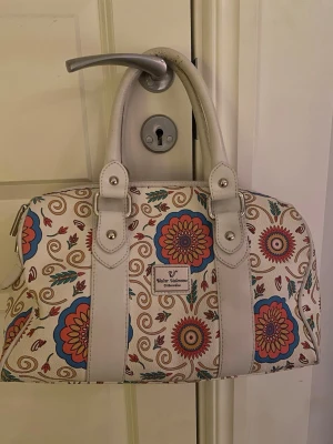 Blommig handväska från Walter Valentino - Säljer en snygg handväska från Walter Valentino med blommigt mönster i färger som röd, blå, beige och turkos. Väskan har vita handtag och detaljer, samt en metallbricka framtill. Materialet är skinnimitation och insidan är fodrad med satin. Perfekt storlek för allt du behöver.