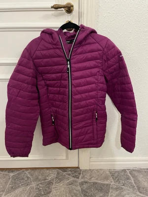 Mörklila quiltad pufferjacka Luxer - Quiltad pufferjacka från Luxer i mörklila med huva och dragkedja framtill. Jackan har två fickor med dragkedja och ett stilrent, sportigt uttryck. Perfekt för kyliga dagar och enkel att matcha med jeans eller sneakers.