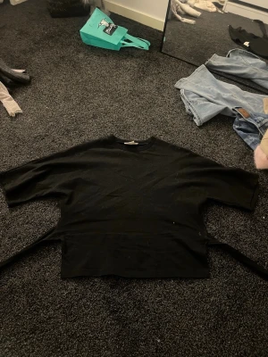 Svart omlottopp från Nelly - Säljer en svart omlottopp från Nelly i storlek XXS. Toppen har korta ärmar och knytband i midjan som ger en snygg omlott-effekt både fram och bak. Materialet är mjukt och känns som bomull. Perfekt att styla med jeans eller kjol för en cool look.