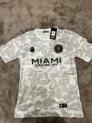 Gråvit camo fotbollströja BAPE x Miami - Limited edition fotbollströja från A Bathing Ape och Inter Miami med gråvit camouflage-mönster, svart MIAMI-tryck och klubbmärke på bröstet. Tröjan har rund hals, korta ärmar och detaljer i rosa och svart. Perfekt för dig som gillar streetwear och fotboll.