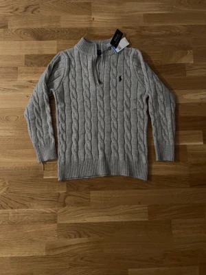  half zip tröja Polo Ralph Lauren - Säljer en grå stickad tröja från Polo Ralph Lauren med klassiskt kabelmönster och half zip framtill. Tröjan har långärmad passform, ribbade muddar och svart broderad logga på bröstet. Perfekt för dig som gillar stilren och tidlös design. Helt ny nästan aldrig använd. Storlek M, men passar mer som en S. 
