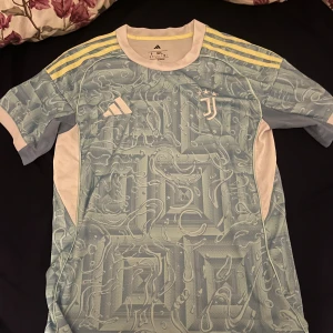 Adidas Juventus fotbollströja blå - Säljer en Juventus fotbollströja från Adidas i ljusblå med mönstrad design och vita detaljer. Tröjan har gula ränder på axlarna och Juventus-logga på bröstet. Perfekt för dig som gillar snygga matchtröjor med unik look. Kan funka som M oxå