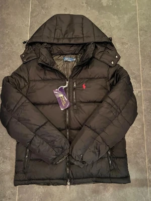 Svart pufferjacka från Polo Ralph Lauren - Svart pufferjacka från Polo Ralph Lauren med huva och dragkedja framtill. Jackan har ett rött broderat Polo-logo på bröstet och flera praktiska fickor med dragkedja. Materialet är polyester och jackan har quiltad design för extra värme.