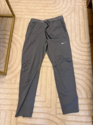 Grå träningsbyxor från Nike - Säljer ett par grå Nike träningsbyxor med snörning i midjan och dragkedjor vid fickorna. Byxorna har en slim passform och är tillverkade i ett lätt syntetmaterial som andas. Perfekta för löpning eller gymmet, med Nike-logga på benet.