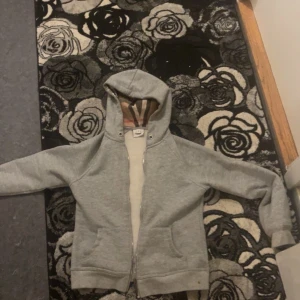 Grå hoodie från Burberry med dragkedja - Säljer en grå hoodie från Burberry i storlek S. Klassisk modell med dragkedja, huva och fickor framtill. Insidan av huvan har det ikoniska Burberry-rutiga mönstret i beige, rött och svart. Perfekt för dig som gillar stilrena och exklusiva plagg.