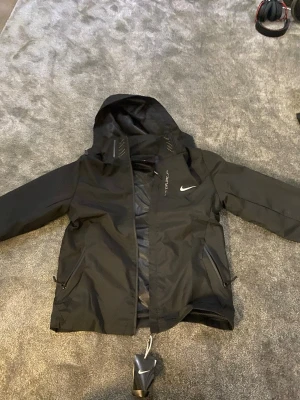 Svart Nike vindjacka med huva - Svart vindjacka från Nike med huva och dragkedja framtill. Jackan har två sidofickor med dragkedja och ett stilrent Nike-logo på bröstet. Materialet är lätt och syntetiskt, perfekt för blåsiga dagar. Snygg och sportig look med clean design.