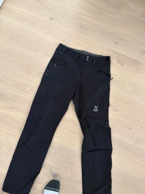 Svarta täckbyxor från Haglöfs storlek m - Svarta täckbyxor från Haglöfs i storlek M. Byxorna har dragkedjor på fickorna och bälteshällor i midjan. Materialet är slitstarkt och passar perfekt för utomhusaktiviteter under kyliga dagar. Snygg, enkel design med diskret Haglöfs-logga på benet.