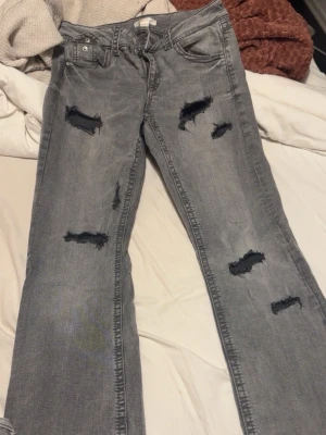 Grå jeans från Gina Tricot - Säljer ett par grå jeans från Gina Tricot med coola slitningar och trasiga detaljer på benen. Modellen har raka ben, klassiska fickor både fram och bak samt knappstängning. Perfekt för dig som gillar en edgy streetstil.
