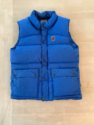 Blå dunväst från Fjällräven - Säljer denna blå dunväst från Fjällräven med hög krage och två stora fickor framtill. Västen har tryckknappar och klassisk Fjällräven-logga på bröstet. Perfekt att slänga över en hoodie för extra värme.