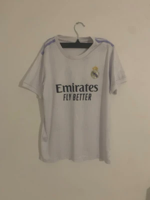 Real Madrid matchtröja Benzema och shorts - Säljer en vit Real Madrid matchtröja och shorts med lila detaljer på axlarna, klubbmärke på bröstet och 'Emirates Fly Better' tryck. På ryggen står det Benzema och nummer 9 i svart. Perfekt för fotbollsfans!