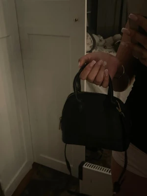 Svart handväska med gulddetaljer - Säljer en stilren svart handväska med rundad form och två handtag. Väskan har guldfärgade dragkedjor och en avtagbar axelrem, dragkedja är trasig och går inte och stänga. Perfekt storlek för det viktigaste och passar till många olika outfits.