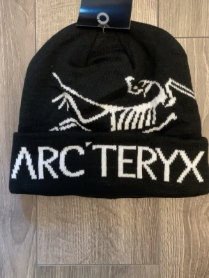 Svart Arc'teryx mössa med logga - Snygg svart mössa från Arc'teryx med vit stickad logga och fossilmotiv framtill. Mössan är ribbstickad och har uppvikt kant för extra stilpoäng. Perfekt för kalla dagar och outdoor-häng.