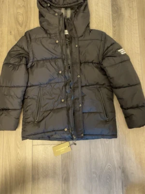 Svart pufferjacka från Burberry - Säljer en svart pufferjacka från Burberry med huva och tydlig logga på ärmen. Jackan har dragkedja framtill, två fickor med dragkedja och quiltad design. Perfekt för kalla dagar och riktigt snygg streetstil.