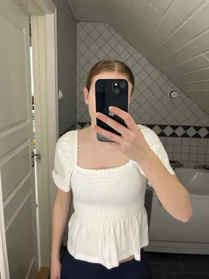 Vit blus från H&M - Supersöt vit blus från H&M med smockad överdel och volang nertill. Blusen har puffiga korta ärmar och fyrkantig ringning både fram och bak. Den är använd några gånger, men är i bra skick. 