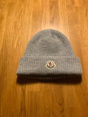 Grå stickad Moncler mössa - Säljer en grå stickad mössa från Moncler med klassisk ribbad design och uppvikt kant. Mössan har Moncler-logga framtill och är tillverkad i mjukt material som håller dig varm. Perfekt för dig som vill ha en clean och stilren look.