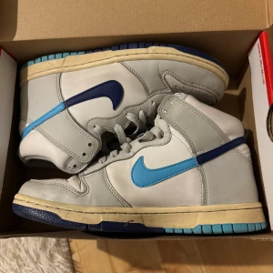 Nike Dunk High vit/blå sneakers - Nike Dunk High sneakers i vitt och olika blå nyanser. Skorna har högt skaft, klassisk snörning och den ikoniska swoosh-loggan i blått och ljusblått på sidorna. Ovandelen är i skinn och sulan har blå och beige detaljer. Perfekta för dig som gillar retrostil.