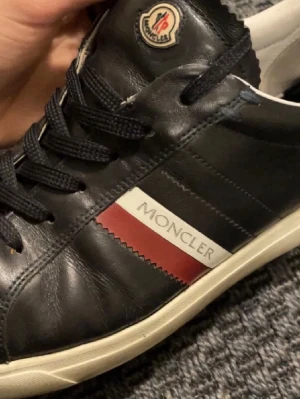 Svarta Moncler sneakers i skinn - Snygga svarta sneakers från Moncler med klassisk röd och vit detalj på sidan och logotyp på plösen. Skorna har snörning och en platt vit sula. Tillverkade i skinn med en rund tå och stilren design som passar till många outfits.