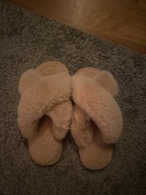 Fluffiga tofflor  - Supermysiga tofflor i beige/ljusrosa med korslagda band och fluffig pälsimitaion. Platt sula och mjuk insida som gör dem sköna att glida runt i hemma. Sitter även bra på foten och faller ej av! Typ aldrig använt💓