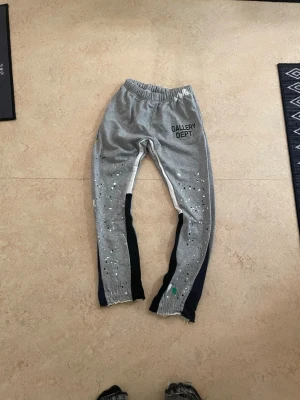 Grå mjukisbyxor från Gallery Dept. - Säljer ett par grå mjukisbyxor från Gallery Dept. med elastisk midja och coolt stänkmönster i vitt och svart på benen. Byxorna har svarta och vita paneler längs sidorna och råa kanter vid bensluten. Perfekt för en avslappnad och trendig look.