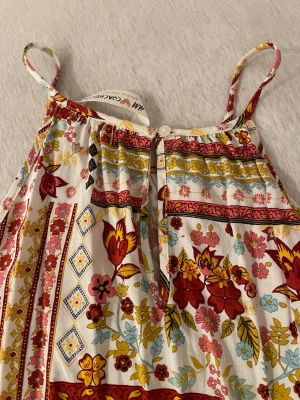 Blommig linnetopp från H&M - Säljer en färgglad linnetopp från H&M med blommigt mönster i rött, gult, rosa och blått på vit botten. Toppen har smala axelband, knappdetalj framtill och dekorativt band med små tofsar längst ner. Perfekt för varma dagar!
