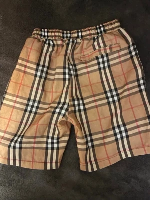 Beige rutiga shorts från Burberry - Säljer ett par klassiska beige shorts från Burberry med det ikoniska rutiga mönstret i svart, vitt och rött. De har elastisk midja och fickor både fram och bak. Perfekta för dig som vill ha en snygg och tidlös look.
