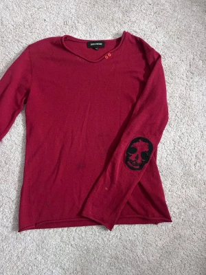 Vinröd långärmad tröja Zadig & Voltaire - Vinröd långärmad tröja från Zadig & Voltaire med två röda knappar vid halsringningen och svarta döskalletryck på ärmarna. Tröjan är tunnstickad och har diskret ZV-detalj nertill. Perfekt för dig som gillar edgy detaljer och stilren design.