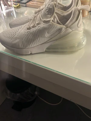 Nike Air Max 270 sneakers vita - Nike Air Max 270 sneakers i helvit färg med mesh-överdel och stor synlig Air Max-sula. Skorna har snörning, rund tå och platt sula. Snyggt och sportigt utseende med diskreta Nike-loggor på sidan och tungan. Perfekt för dig som gillar stilrena och bekväma sneakers.