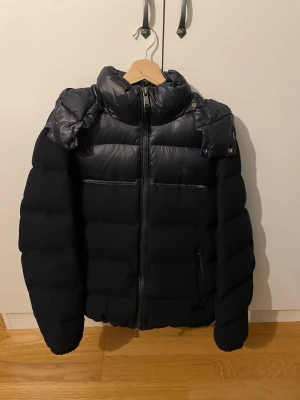 Marinblå pufferjacka Polo Ralph Lauren - Säljer en marinblå pufferjacka från Polo Ralph Lauren med glansiga paneler upptill och på huvan. Jackan har dragkedja framtill, två sidofickor med dragkedja och en hög krage. Materialet är en mix av polyester och dun, perfekt för kalla dagar.