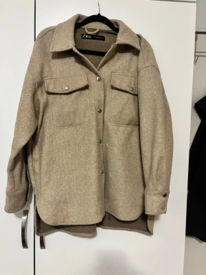 Beige overshirt från Zara - Säljer en beige overshirt från Zara med klassisk krage, stora bröstfickor med knappar och knappstängning framtill. Jackan har långärmade ärmar med knappdetaljer vid mudden och är tillverkad i ett mjukt ullmaterial. Perfekt för lager-på-lager-stil.