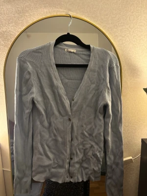 Ljusblå ribbad kofta från Gina Tricot - Säljer en ljusblå ribbad kofta från Gina Tricot med v-ringning och knappar framtill. Koftan har långärmad design och är tillverkad i mjuk bomullsmix som är skön att ha på sig. Perfekt att slänga över en topp för en chill look.