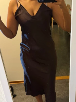 Brun satin klänning från Zara - Säljer en brun långklänning från Zara i glansigt satinmaterial. Klänningen har v-ringad hals, smala axelband och en figurnära passform som ger en snygg siluett. Perfekt för dig som gillar stilrena och trendiga plagg.