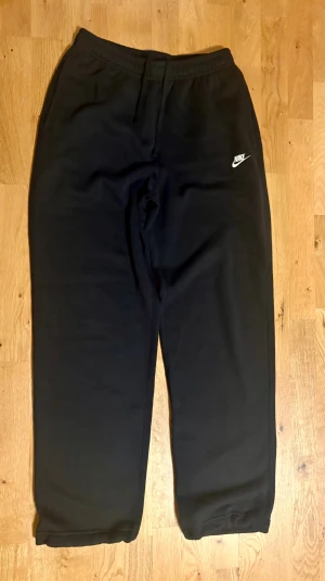 Svarta mjukisbyxor från Nike, storlek S - Svarta mjukisbyxor från Nike med klassisk logga på vänster lår. Byxorna har elastisk midja med snörning, bakficka med knapp och justerbara benslut. Tillverkade i mjukt bomullsmaterial som är skönt mot huden. Perfekta för chill eller träning.