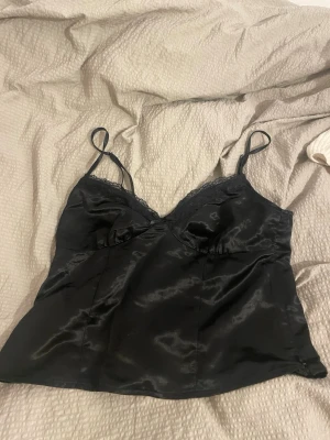 Svart satin linnetopp från H&M Divided - Säljer en svart linnetopp från H&M Divided i storlek 36. Toppen är gjord i glansigt satinmaterial med tunna axelband och spetsdetaljer vid bysten. Perfekt att styla med jeans eller kjol för en cool look.