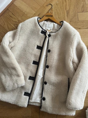 Vit teddyjacka med svarta detaljer - Säljer en fluffig vit teddyjacka från H&M med svarta knappar och svarta kantdetaljer. Jackan har rund hals, lång ärm och två fickor framtill. Insidan är fodrad i ljus satin och jackan har en loose passform som är perfekt för kyliga dagar.