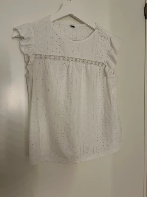 Vit blus med spets och volang - Säljer en söt vit blus med broderade hålmönster och volangdetaljer på axlarna. Blusen har rund hals och en dekorativ spetsrad framtill. Perfekt för sommarens alla tillfällen, lätt och luftig i bomull.