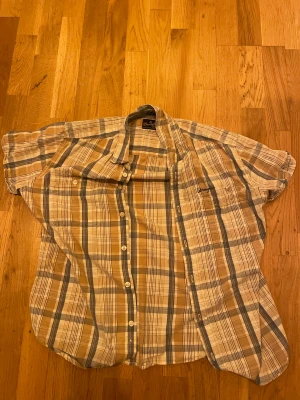 Rutig kortärmad skjorta från Wrangler - Säljer en rutig skjorta från Wrangler i beige, blå och vit. Skjortan har korta ärmar, klassisk krage och knappar framtill. Perfekt för dig som gillar avslappnad stil och vill ha något lätt och luftigt. Materialet är mjuk bomull som känns skönt mot huden.